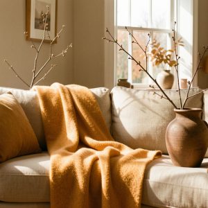 Salon lumineux décoré pour l’automne, avec un canapé beige, un plaid en laine ocre, un vase en grès brun et quelques branchages.