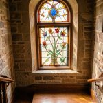 Quel vitrail choisir pour la fenêtre d’escalier d’une maison ancienne ? Vitrail aux motifs botaniques et géométriques installé dans la fenêtre d’un escalier de maison ancienne, éclairé par une douce lumière matinale.