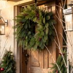 Quelles décorations choisir pour un porche de Noël 2026 durable et économique ? Porche d'une maison française décoré pour Noël avec une couronne de pin, des lanternes métalliques, des branches de bouleau et des tons bordeaux et vert mousse dans une lumière chaude de fin d'après-midi.