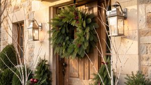 Porche d'une maison française décoré pour Noël avec une couronne de pin, des lanternes métalliques, des branches de bouleau et des tons bordeaux et vert mousse dans une lumière chaude de fin d'après-midi.